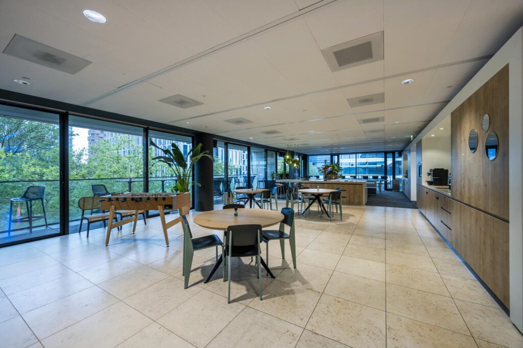 Modern office lounge on Gustav Mahlerlaan with round tables, a foosball table, and large windows overlooking greenery.